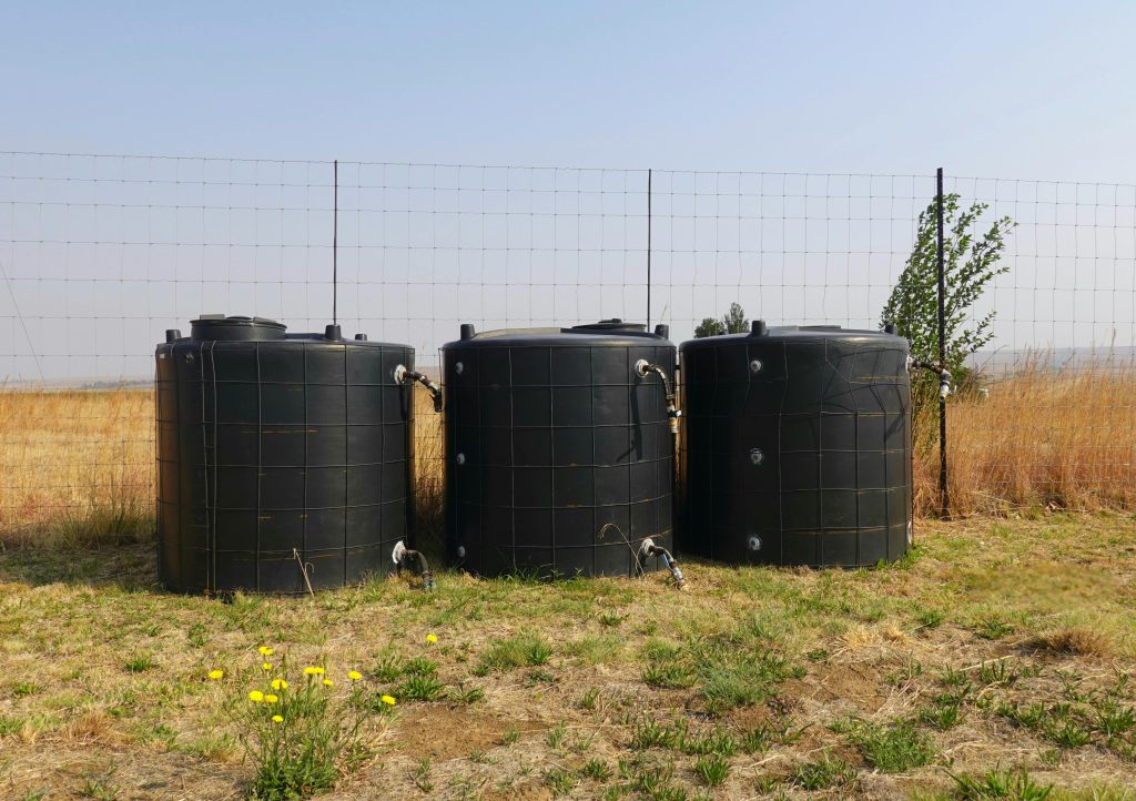 Three black water tanks in a dry, grassy field with a wire fence.