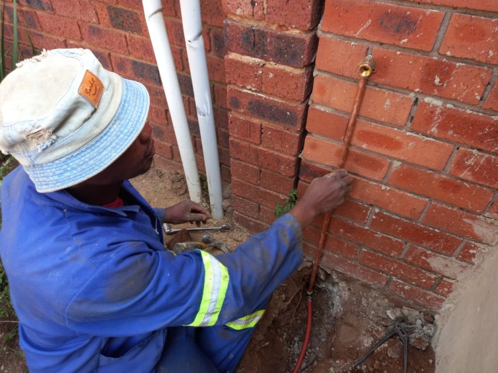 A plumber in blue overalls installing a vertical copper water pipe and brass fitting onto an outdoor red brick wall