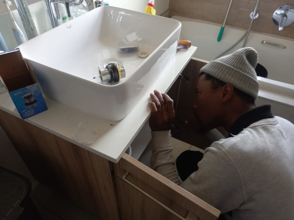 A plumber looking inside a wooden vanity cabinet to install a new chrome pop up drain assembly into a white rectangular ceramic sink basin