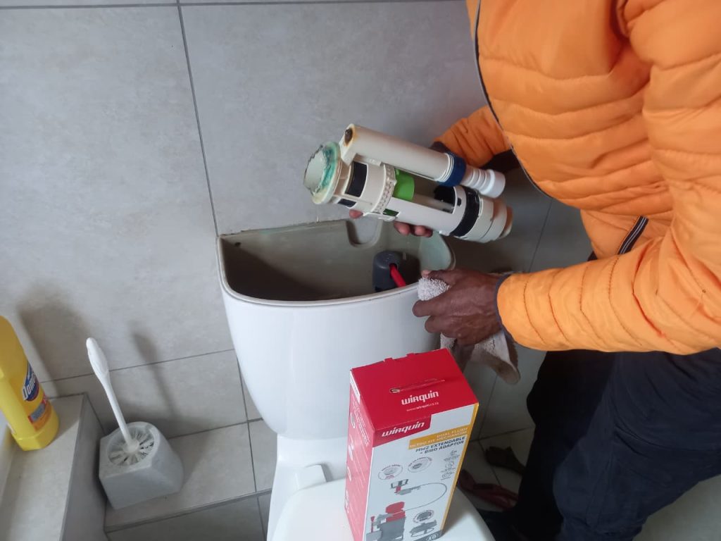 a plumber holding a an old toilet flush valve over an open ceramic cistern next to a red wirquin parts box